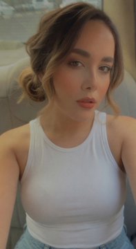 A close-up selfie of a young woman with brown hair wearing a white tank top, with the image stretched vertically to exaggerate her proportions.