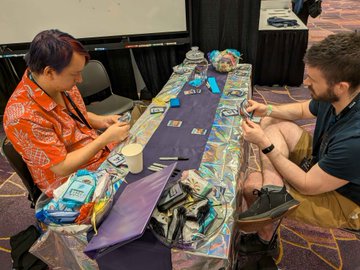 Two people sit at a #RareEvo booth exchanging real-life trading cards across a colorful table display, exemplifying the ecosystem-building activities highlighted in the post.