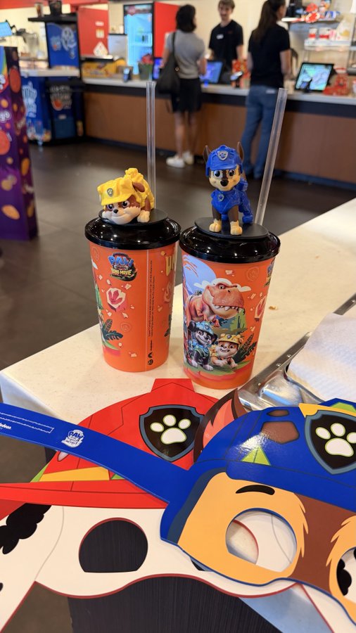 Two PAW Patrol themed cups with character toppers (Rubble and Chase) sit on a table at what appears to be an AMC movie theater concession area, accompanied by PAW Patrol themed masks and merchandise.

The image relates to the "#atAMC $AMC" post as it captures a visit to an AMC movie theater, showing branded promotional kids' meal items available at the concession stand.