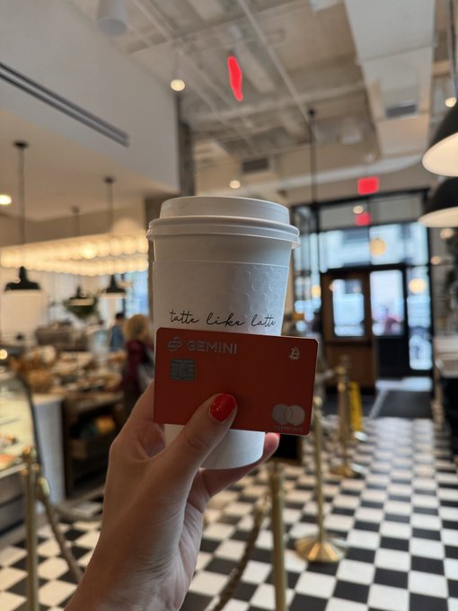 A hand holds a coffee cup labeled 'tatte like latte' alongside a red Gemini bank card in a brightly lit café with a checkered floor.