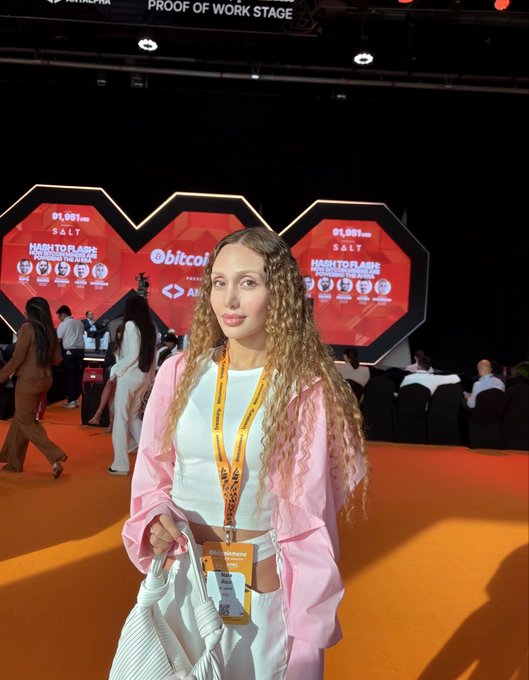 A young woman with long curly blonde hair wearing a pink jacket and lanyard stands at an event. The stage displays 'Proof of Work Stage' and 'bitcoin' with signs showing '₿1,991,406 SALT'.