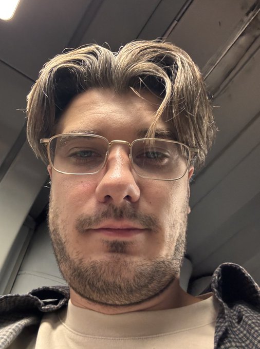 A close-up portrait of a man with brown hair and a light beard, wearing wire-rimmed glasses and a plaid shirt over a beige t-shirt, sitting inside an airplane.