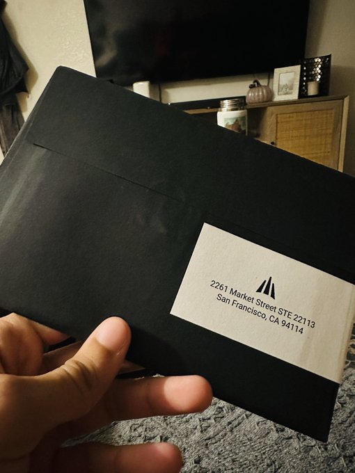 A hand holds up a black envelope with a white label addressed to '2261 Market Street STE 221113, San Francisco, CA 94114' featuring a small triangular logo.