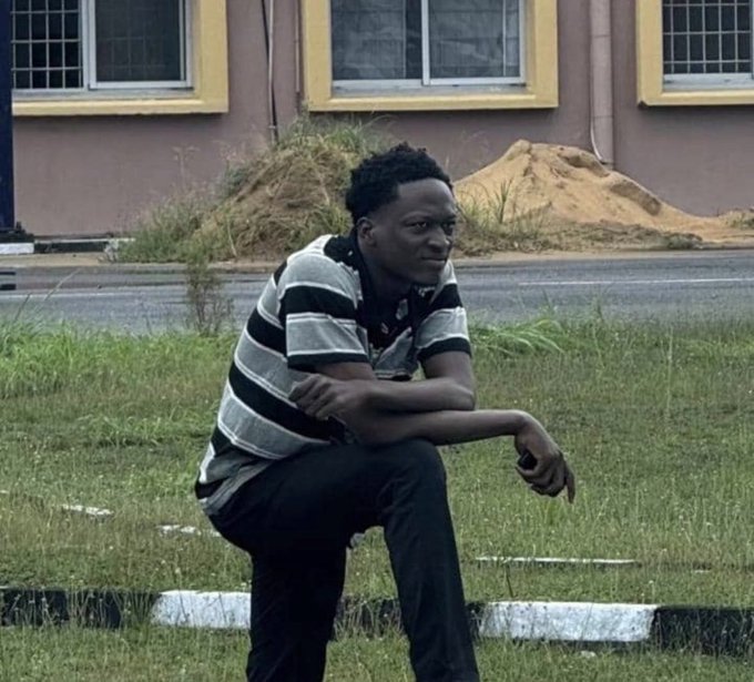 A young man wearing a striped polo shirt and dark pants crouches on a grassy patch next to a road, looking sideways at the camera, with a building in the background.