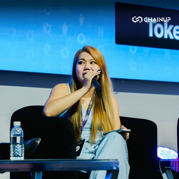 A photograph of a woman with long brown hair speaking into a microphone at what appears to be a ChainUp event, with a partially visible 'Token' sign in the background.