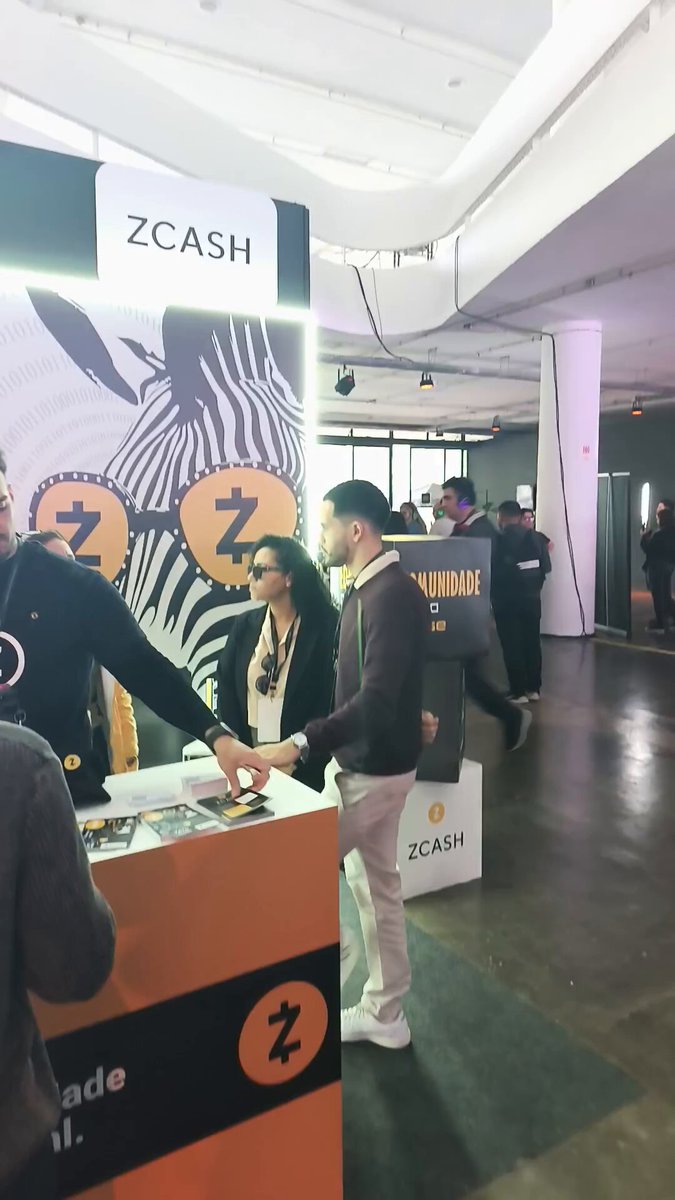 The image shows a group of people standing around an event booth for Zcash, which has a zebra design on its side. The booth appears to be located inside a building with white walls and a ceiling that features a curved line. There are several people in the scene, some of whom appear to be engaged in conversation or examining the booth's offerings. One person is holding a cell phone, possibly taking a photo or recording information about the event.