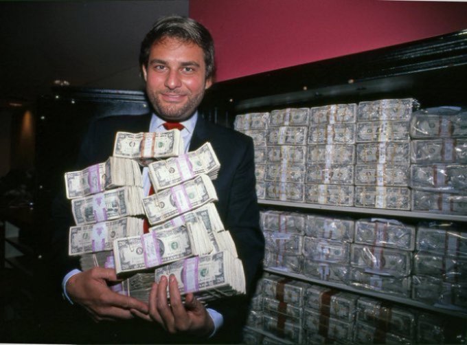 A man in a dark suit and red tie proudly holds a fan of $20 U.S. bills in front of stacked currency bundles, smiling confidently to evoke the victorious "winner" feeling promised by the post.