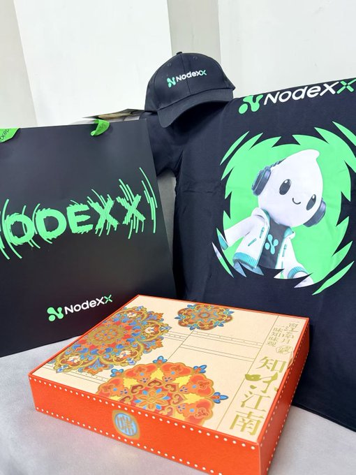 A promotional merchandise package from NodeXX featuring a branded black cap, a t-shirt with a green character design, and an ornately decorated traditional box.