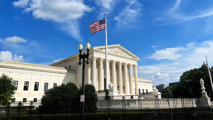 A neoclassical white government building with tall columns, an American flag flying on a flagpole, and an ornate black lamppost in the foreground against a blue sky with scattered clouds.