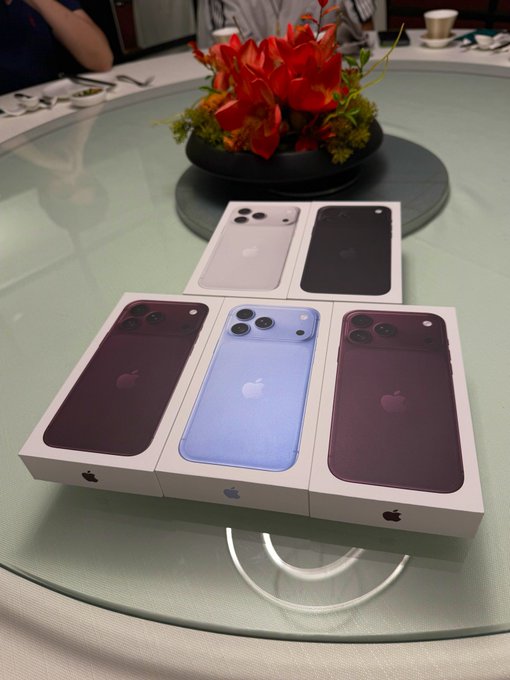 Five boxed Apple iPhones arranged on a glass table, showing various colors including silver, black, dark red, light blue, and another dark red/purple shade, with a floral centerpiece in the background.