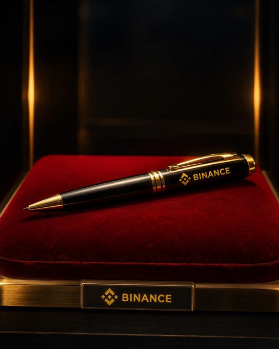 A luxury black and gold pen branded with the 'BINANCE' logo rests on a red velvet cushion inside an elegant display case, with a matching gold plate below also bearing the 'BINANCE' name.