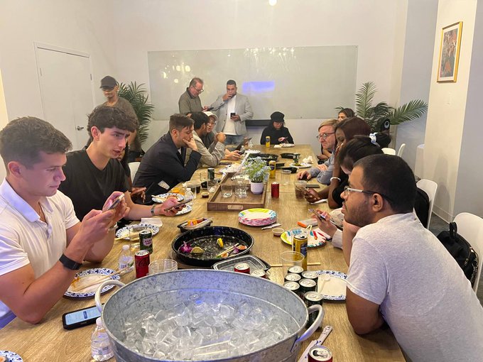 A diverse group of people sit around a long wooden table sharing a meal with drinks, sushi plates, and a metal ice bucket in the foreground.