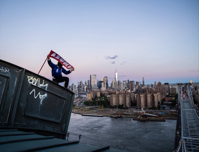 A person crouches atop a graffiti-marked rooftop structure, triumphantly holding up an "AZUKI" banner against the backdrop of the Manhattan skyline and Brooklyn Bridge at dusk, capturing the urban exploration and brand promotion aesthetic associated with the Azuki NFT collection.