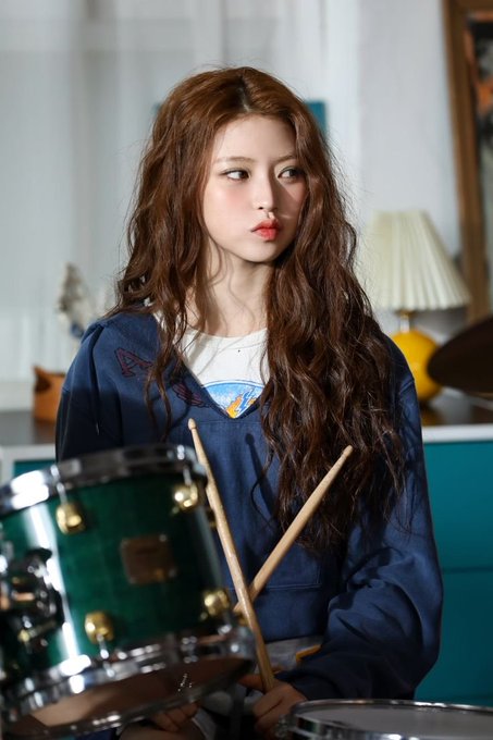 A young woman with long curly brown hair and pink lips holding drumsticks behind a green snare drum, wearing a dark blue jacket over a graphic t-shirt.