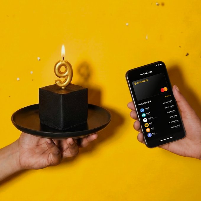 The image features a hand holding a black plate with a black cube-shaped cake topped with a lit gold "9" candle, while another hand holds a smartphone displaying the Binance card app screen — visually celebrating Binance's 9th anniversary and promoting the crypto-powered payment card, as mentioned in the accompanying post.

Key text visible: "MI TARJETA," "BINANCE," "PAGAR CON," along with cryptocurrency options like USDC, USDT, BUSD, BNB, BTC, and ETH.