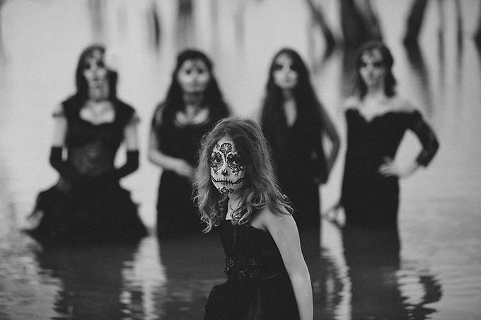 A black-and-white photograph depicts a young girl with intricate skull face paint standing in water, while four blurred figures in dark dresses stand behind her.