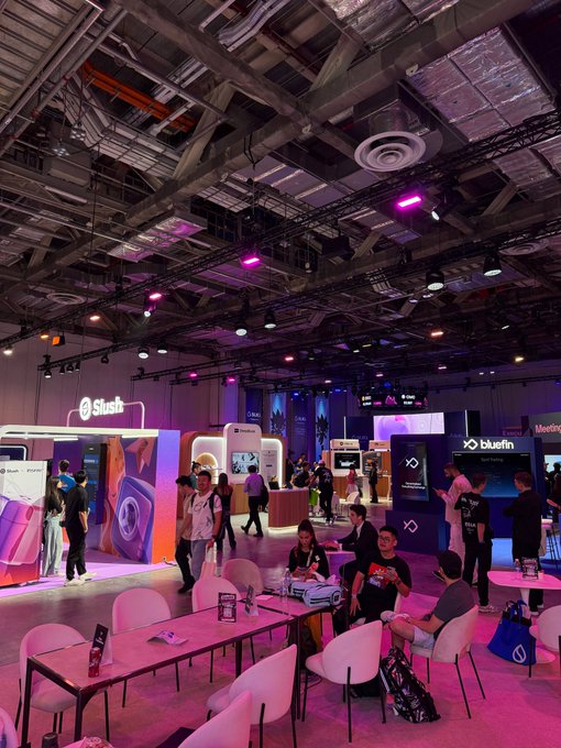 The Whitelist team attends Sui Basecamp 2026 at Marina Bay Sands in Singapore, where they explore a vibrant expo floor featuring booths for sponsors like Sui, Bluefin, and others, with attendees networking around tables bathed in purple lighting.