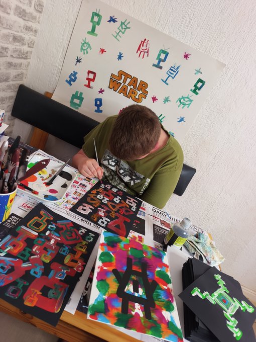 A young boy is seen from above painting Star Wars-themed artwork, with a finished 'Star Wars' poster featuring various character icons displayed behind him.