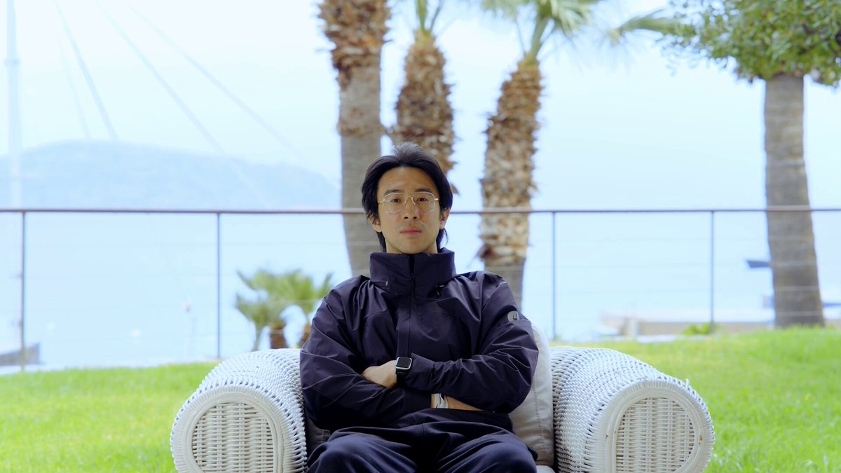 The image features a man sitting in a wicker chair with his arms crossed while wearing glasses. The scene takes place outdoors, possibly at an event or gathering. In the background, there are palm trees and a body of water, creating a relaxing atmosphere. The photo is related to the post about stablecoins as it showcases how people can use them for payments in various situations, including outdoor events where fees might be unpredictable.