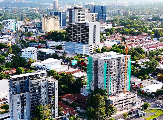 An aerial photograph showing a cityscape with multiple high-rise buildings, including a large structure under construction covered in green netting, surrounded by smaller buildings and palm trees.
