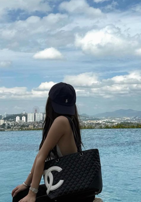 A woman wearing a navy Polo Ralph Lauren cap and holding a black Chanel tote bag sits by a large pool overlooking a cityscape.