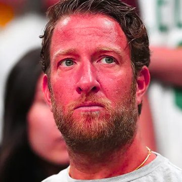 The image shows a man with a beard wearing a grey shirt who appears to be looking off into the distance or deep thought. The background of the photo includes other people present but they are blurred out, making them less noticeable compared to the main subject.