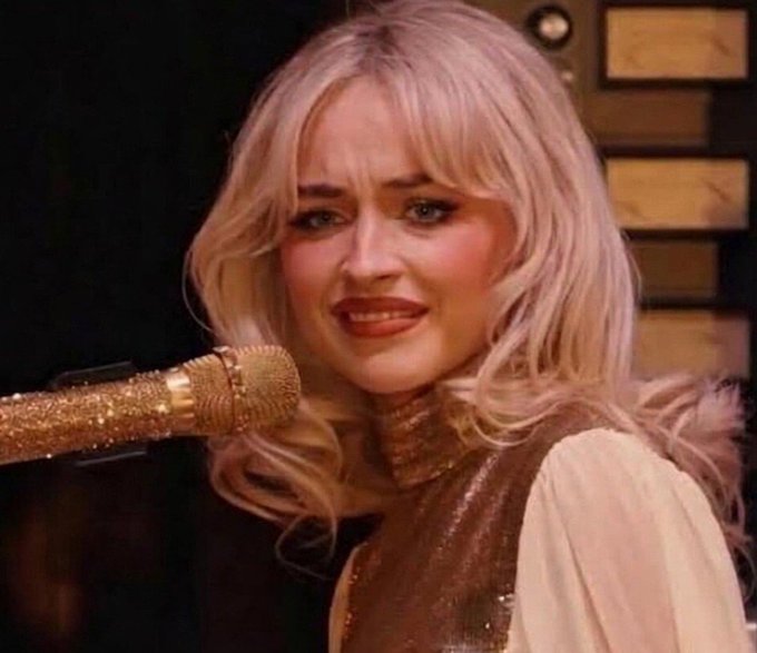 A woman with long blonde hair wearing a tan and gold outfit is smiling while speaking into a sparkly gold microphone, appearing to be on a stage or talk show set.