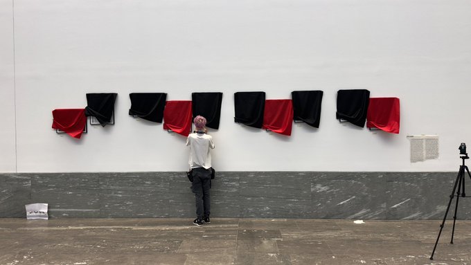A person standing in a gallery looking up at an art installation of alternating red and black cloths mounted on a white wall, with a tripod nearby.