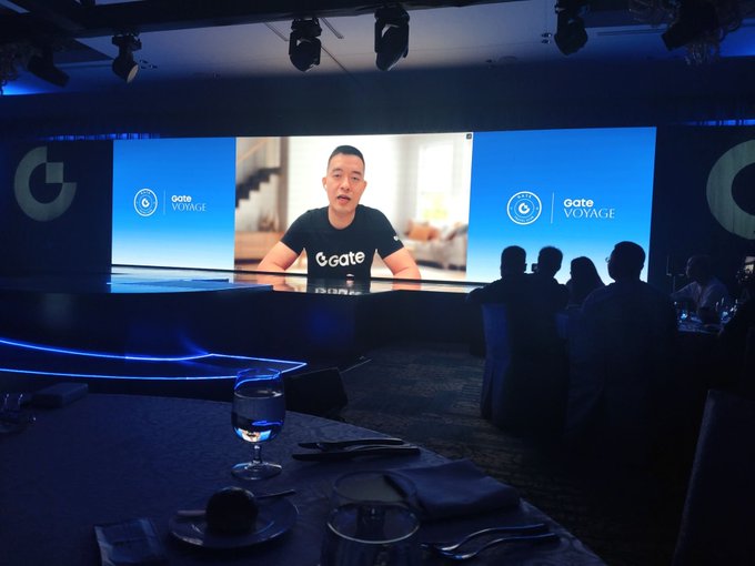 An audience sits at dimly lit tables watching a large screen broadcast a presentation for 'gate VOYAGE', featuring a man in a black 'gate' t-shirt speaking to the camera.