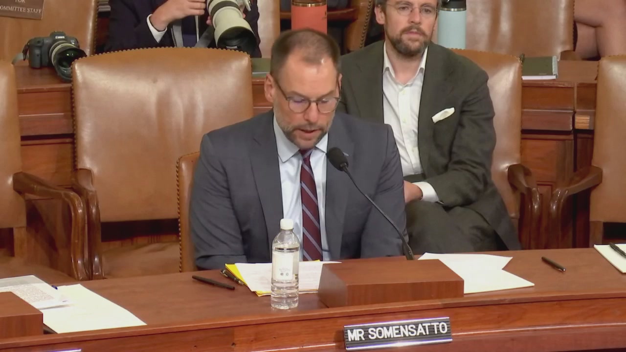 The image shows a man sitting at a wooden desk, wearing glasses and a suit, with another person seated behind him. They appear to be engaged in a discussion or presentation related to Bitcoin and cryptocurrency tax planning. The room has several chairs around the table where they are seated, suggesting that it is likely an official hearing or meeting. A water bottle can also be seen on the desk next to the man speaking.