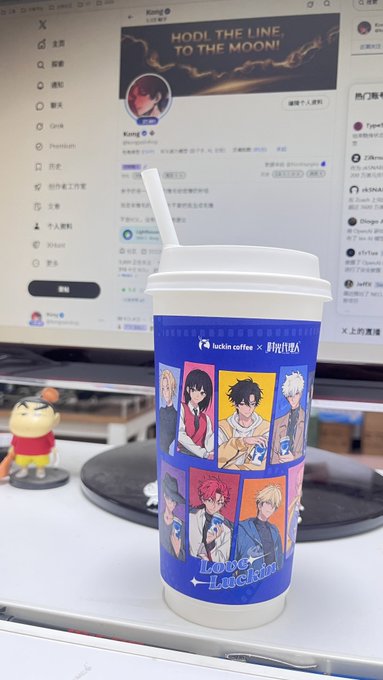 A Luckin Coffee cup featuring anime characters from 'Love and Luckin' collaboration sitting on a desk, with a computer screen in the background displaying a social media profile with the banner 'HODL THE LINE TO THE MOON!'.
