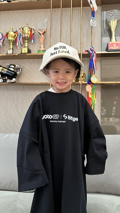 A smiling young child wearing a white bucket hat reading 'No Flu, Just Food' and an oversized black shirt with 'Jotocp | Bitge Regional Partner' printed on the chest, standing in front of a shelf of trophies.