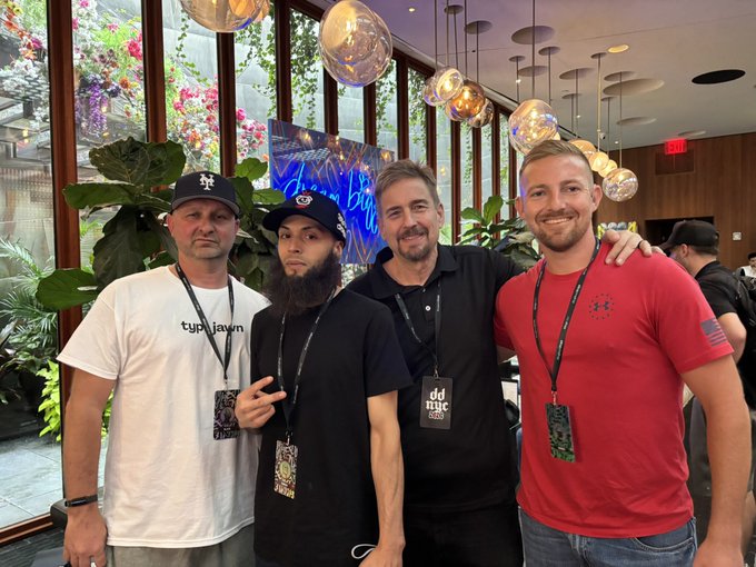 Four men pose together for a photo at an indoor event; they are wearing lanyards with badges, including badges reading 'ddmg', and one man on the left wears a shirt with 'type jawan'.
