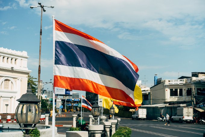 The image shows a large Thai national flag—featuring its distinctive red, white, and blue horizontal stripes—waving prominently in the foreground against a backdrop of urban buildings and a clear blue sky, visually symbolizing the country referenced in the post. A second Thai flag is visible in the background, reinforcing the national context of Thailand's new crypto tax exemption policy that will eliminate capital gains taxes on crypto investments made through SEC-licensed platforms from 2025 to 2029.