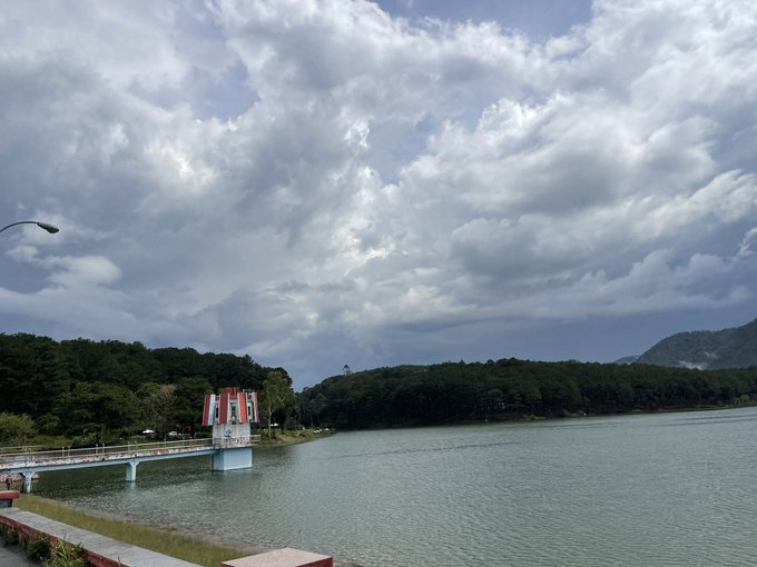 A scenic view of a lake surrounded by forested hills under a cloudy sky, with a small white and red structure on a pier extending into the water.