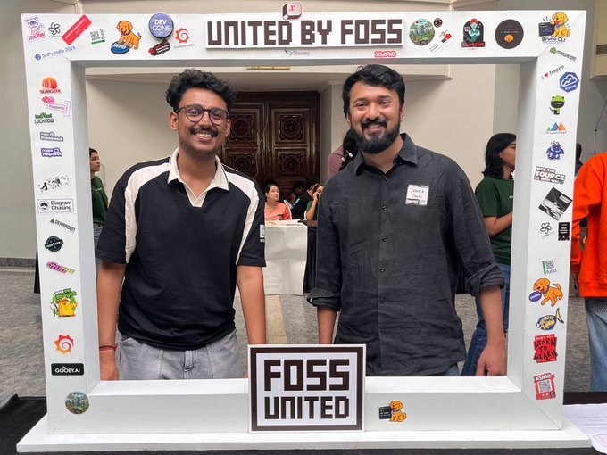 Two smiling men pose inside a 'UNITED BY FOSS' photo booth frame covered in various tech and community stickers at a FOSS United event.