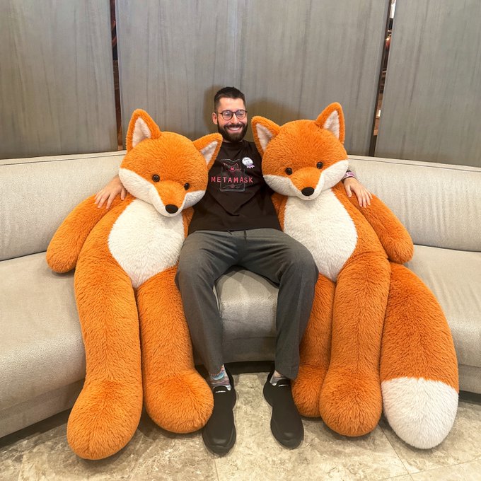 A man sits on a couch flanked by two oversized orange and white fox plush toys, wearing a black 'METAMASK' t-shirt.