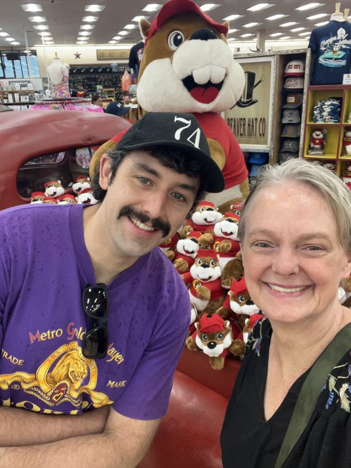 A young man wearing a "74" cap and a purple "Metro Gold" t-shirt takes a selfie with an older woman in front of a large stuffed beaver mascot wearing a red cap, likely at a Trader Hat Co. store filled with merchandise.