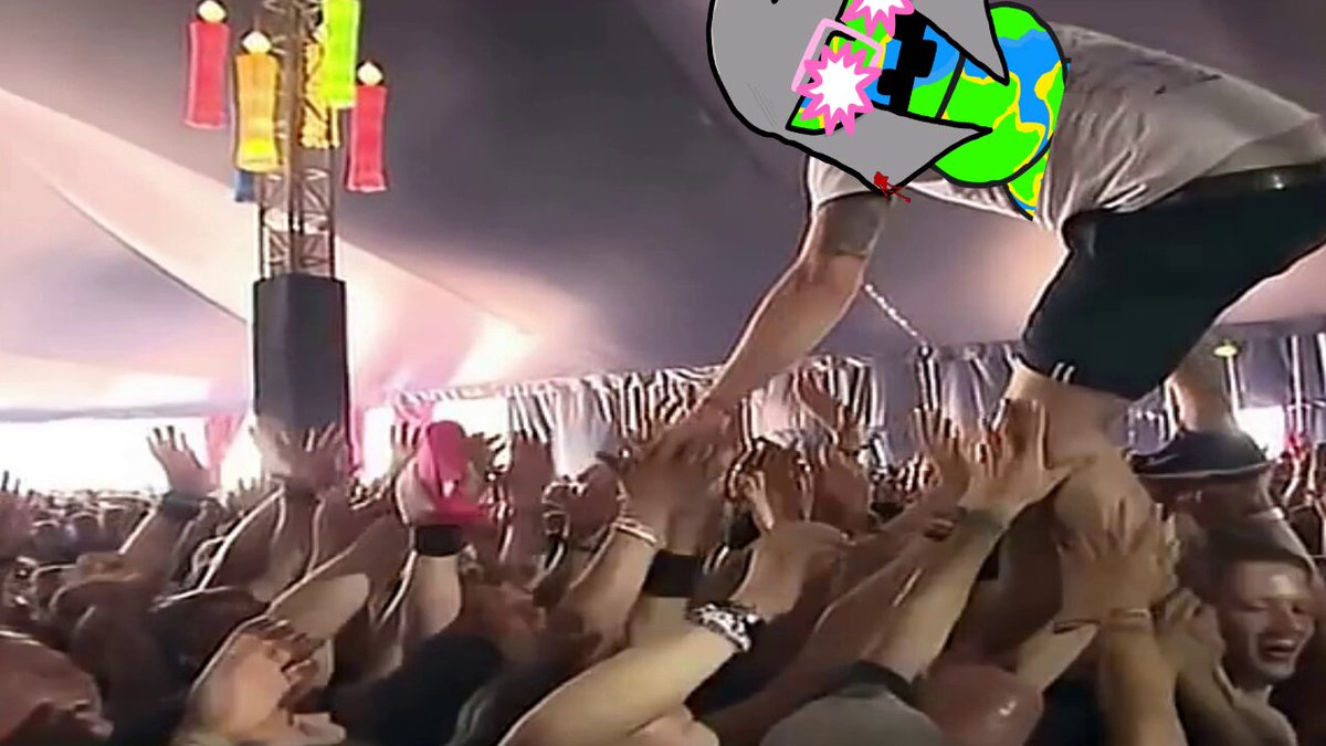 The image shows a large crowd of people gathered together, with some individuals wearing black clothing and others in colorful outfits. A man dressed in black is being lifted into the air by several other people who are holding onto him. The scene appears to be taking place at an outdoor event or festival, as evidenced by the presence of tents and a stage.