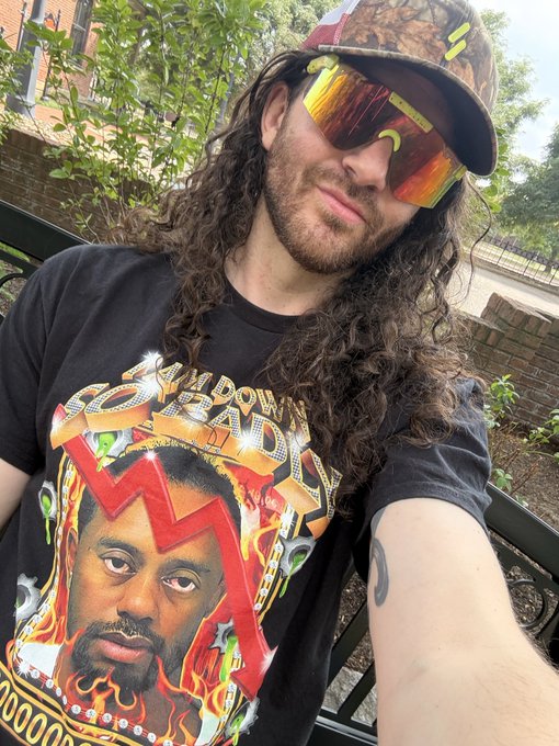 A man with long curly hair, sunglasses, and a camo cap wearing a black graphic t-shirt featuring an artist's portrait, taking a selfie outdoors.