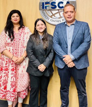 Three people stand posing together in front of an 'IFSCA' (International Financial Services Centres Authority) logo on a wooden wall.