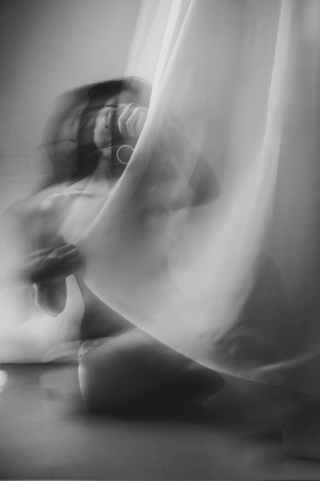 The image shows a woman sitting cross-legged with her legs crossed, partially obscured by a white curtain. The background appears to be a room or studio setting, as there are no other objects visible in the frame. The focus of the photo is on the woman and her position behind the curtain, creating an interesting composition that draws attention to her presence.