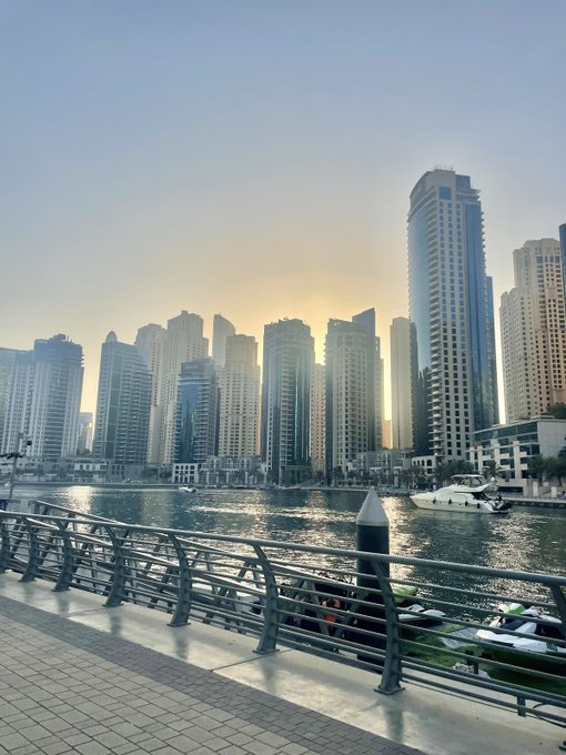 A sunset cityscape showing numerous modern high-rise buildings along a waterfront with boats and yachts docked, viewed from behind a metal railing on a tiled promenade.