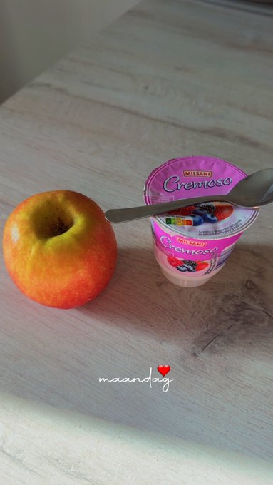 The image features an apple sitting next to a container of yogurt or cottage cheese, both placed on a wooden table. The apple appears to be half-eaten, suggesting that someone has been enjoying its taste. A spoon is also present near the apple, indicating that the person might have used it for eating the fruit. The background of the image seems to be blurred and out of focus, drawing attention to the main subjects in the foreground - the apple and the yogurt container.