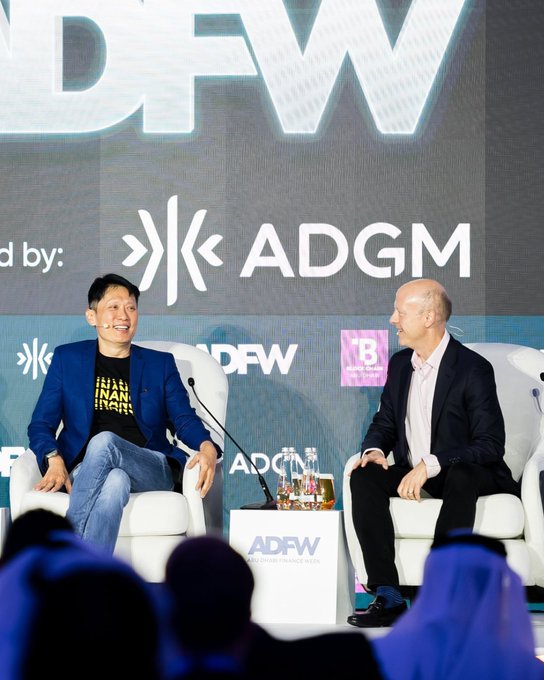 Two men sit on stage at the Abu Dhabi Finance Week (ADFW) event, hosted by ADGM, sharing a lighthearted moment during a panel discussion, with one wearing a Binance shirt. The image visually supports the accompanying post about Binance's $100M commitment to Circle and a five-year partnership extension, highlighting the collaborative spirit behind the deal.