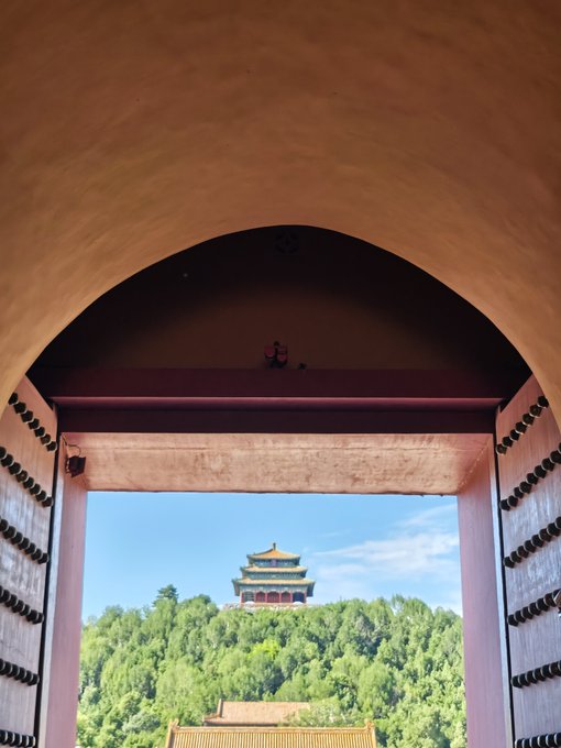 A traditional Chinese gate or pavilion frames a view of a multi-tiered pagoda tower rising above lush green trees against a blue sky.