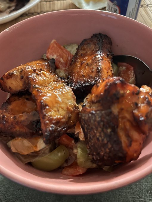 A pink bowl filled with a meal containing grilled or blackened protein pieces (likely chicken or fish), tomatoes, and olives, served over rice.