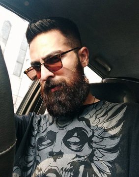 A bearded man wearing tinted sunglasses and a gray graphic t-shirt sits inside a car, looking downward.