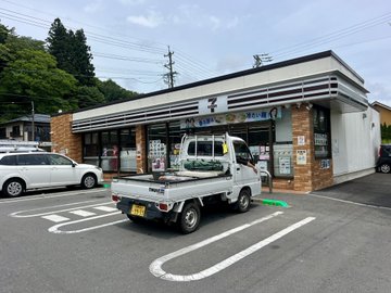 A 7-Eleven convenience store with a Japanese sign reading 'セブン-イレブン' and a small white kei truck parked in front.