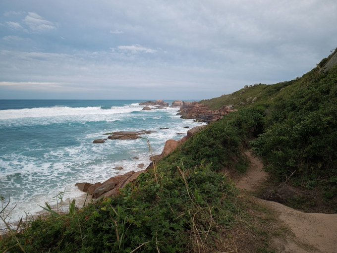 A scenic coastal view shows waves crashing against rocky cliffs along a dirt path lined with green coastal vegetation under an overcast sky.